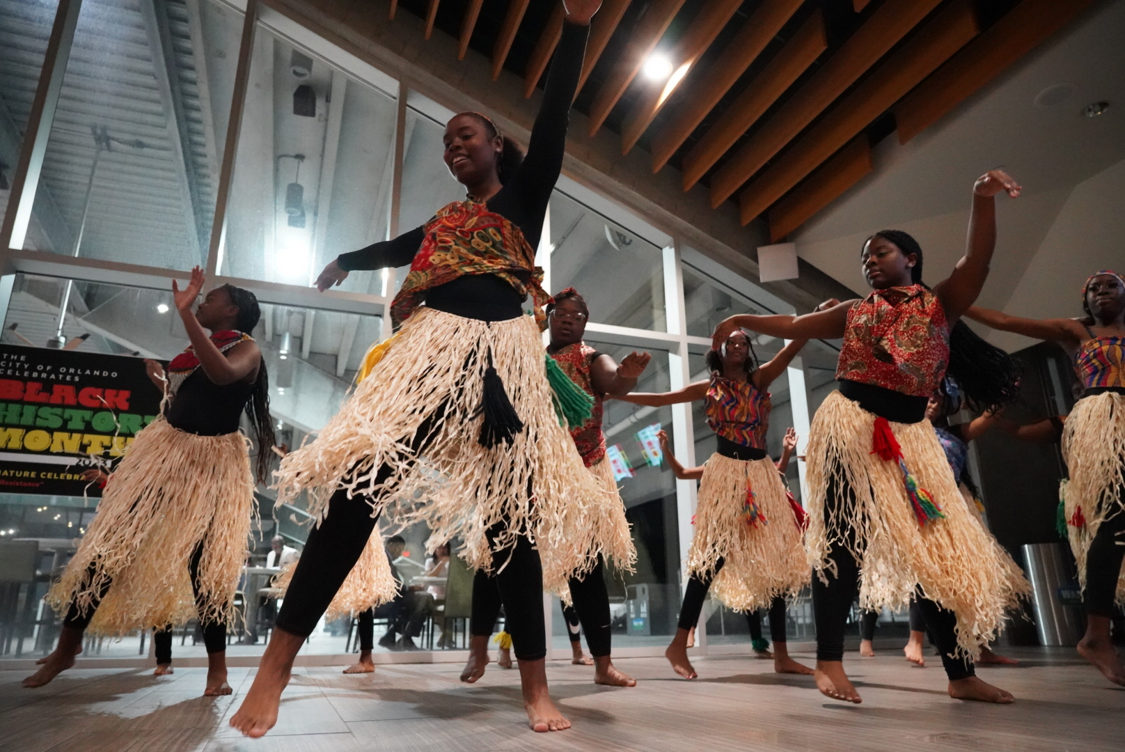 A group of young performers dance in a coordinated formation, wearing traditional African-inspired attire. The attire includes grass skirts and colorful tops. The dance was during a Black History Month celebration at the City of Orlando, highlighting culture, movement, and community.