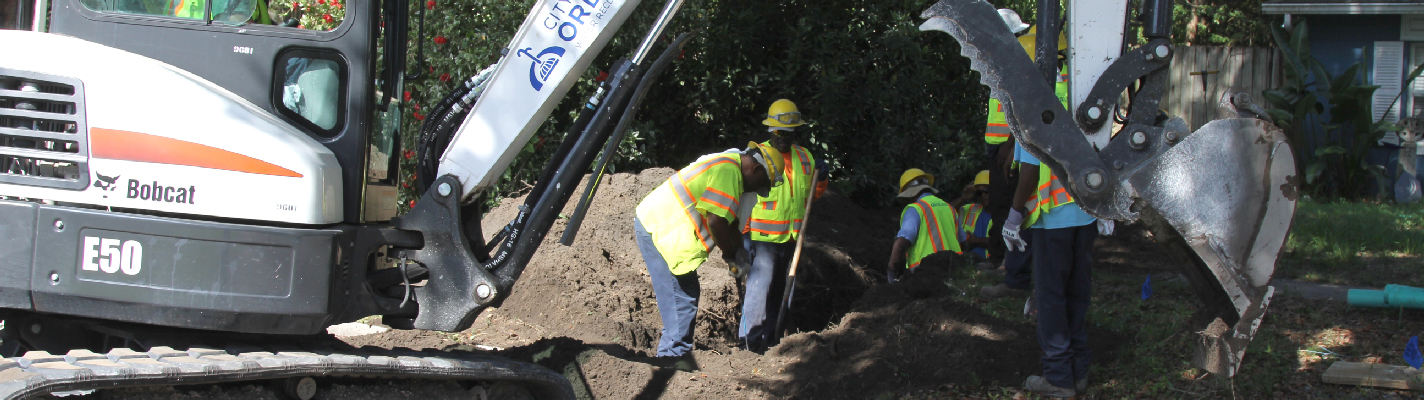 A photo of two City employees wearing personal protective equipment working behind a small white excavator.