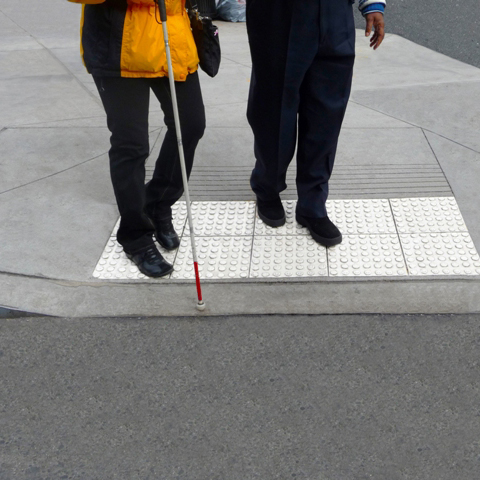 A photo of two pedestrians, one of which is vision disabled, standing on a detectable warning surface on a pedestrian ramp.